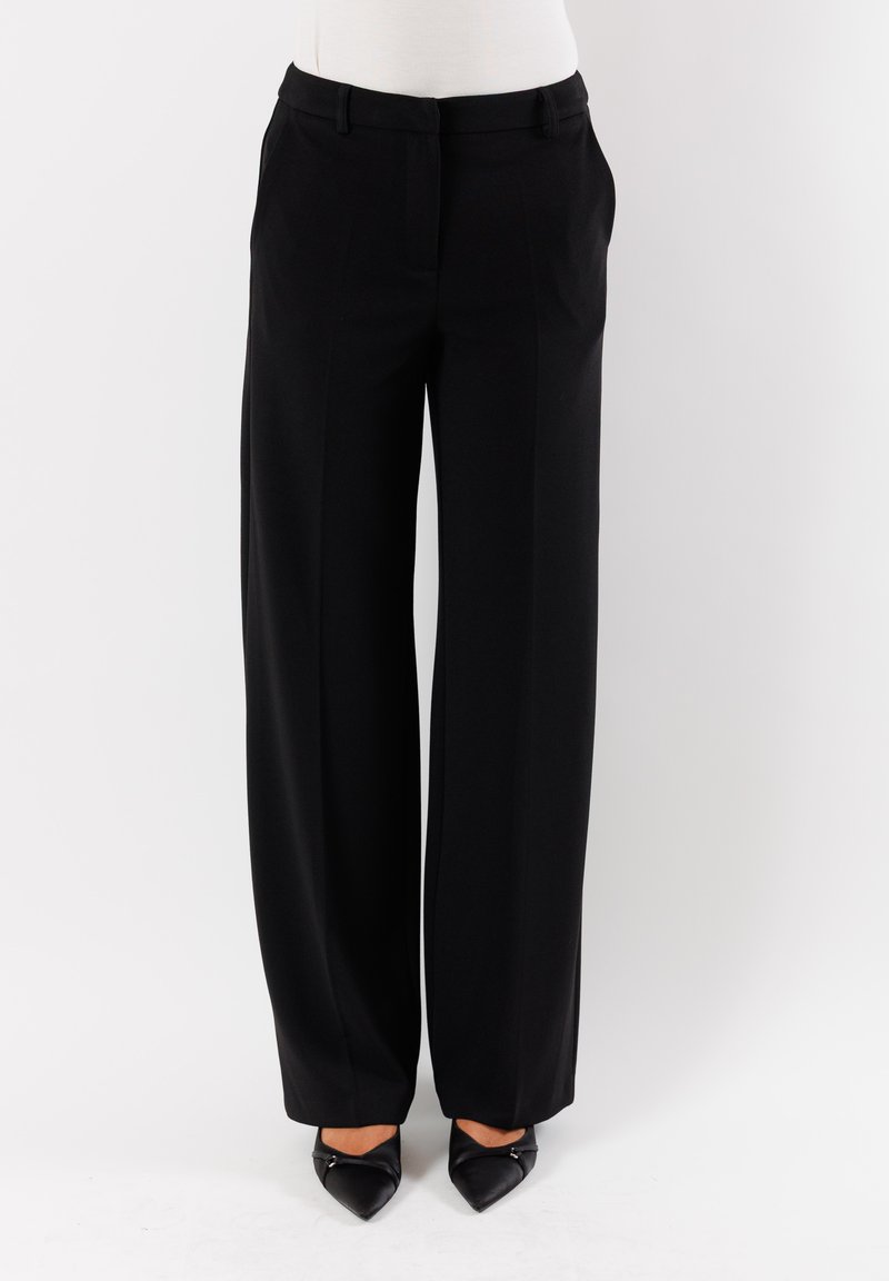 Pantaloni neri a gamba larga realizzati in tessuto morbido con pieghe frontali, tasche laterali e vita. Abbinati a tacchi neri a punta.