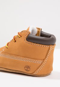 Stivale in suede marrone con lacci gialli e rossi, fodera in shearling bianco e colletto imbottito marrone. Presenta cuciture bianche a contrasto e logo.