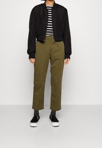 Veste bomber noire, chemise rayée, pantalon vert olive, chaussures noires à enfiler, chaussettes grises à motif. Matières lisses, coupe décontractée, sans embellissements visibles.