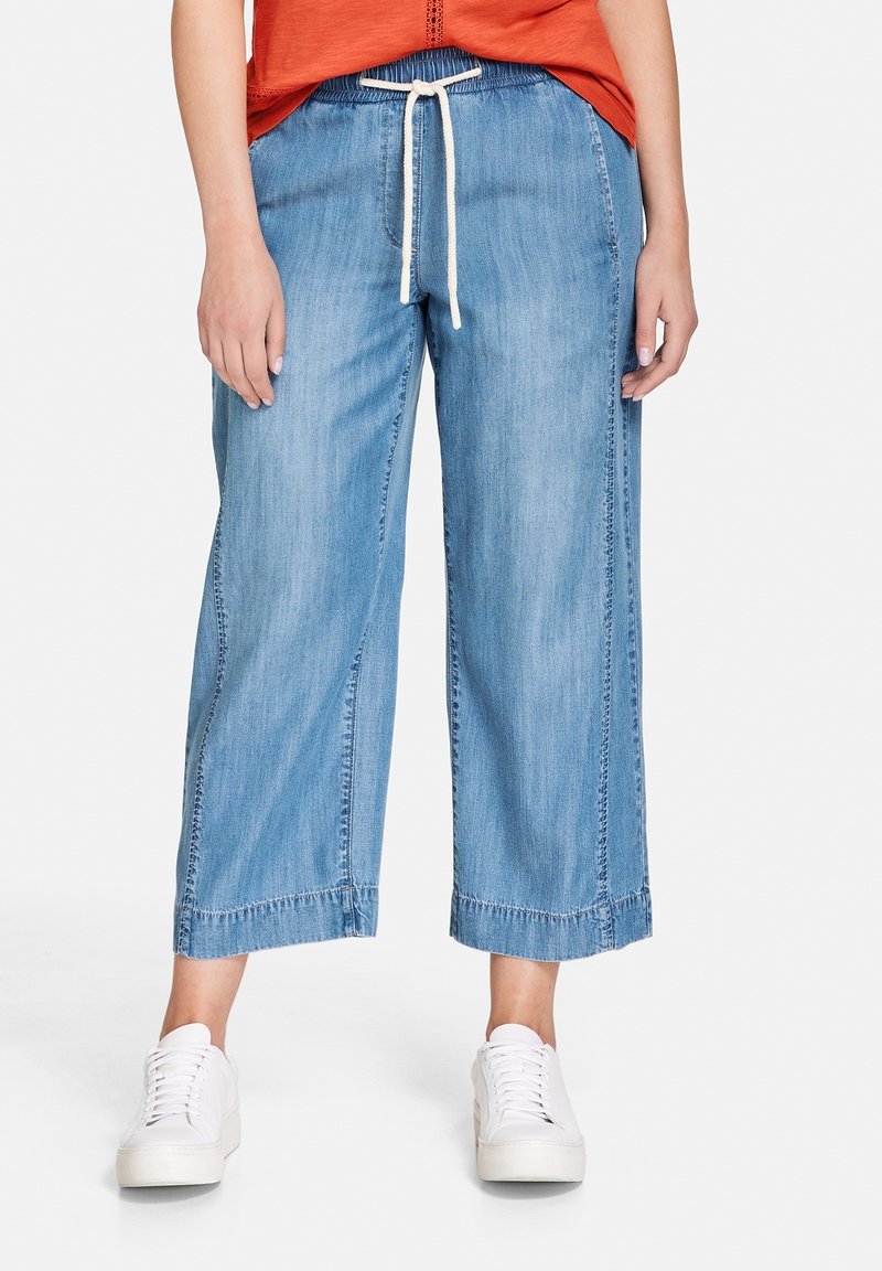 Jean large décontracté bleu clair, raccourci, avec taille à cordon blanc, porté avec des baskets blanches et un haut orange.