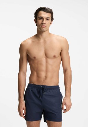 Ung mand med kort mørkt hår iført marineblå badeshorts, stående med et neutralt udtryk foran en ensfarvet hvid baggrund.