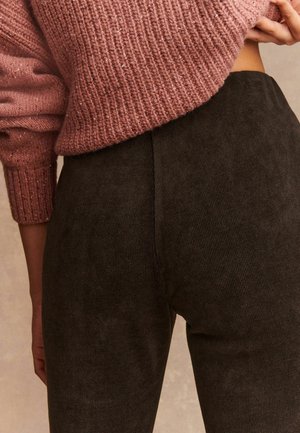 Primo piano di una persona che indossa un maglione lavorato a maglia rosa e pantaloni in velluto a coste marrone scuro, con una mano che solleva leggermente il maglione.