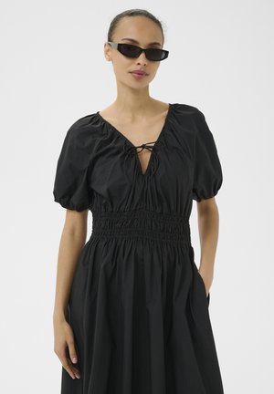 Mujer que lleva un vestido negro con mangas abullonadas, cintura fruncida y lazo en el escote, complementado con gafas de sol negras estrechas, posando con una mano en el bolsillo.