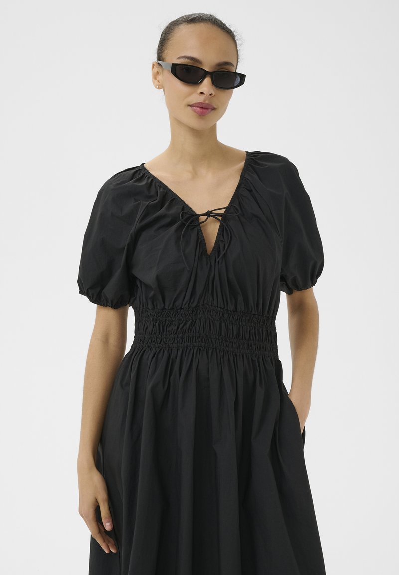 Woman wearing a black puff-sleeve dress with smocked waist and neckline tie, accessorized with narrow black sunglasses, posing with one hand in pocket.