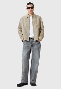 Beige mockaskinnjacka med krage, dragkedjestängning och sidofickor, parat med lösa grå jeans och svarta skor. Modellen bär mörka solglasögon.