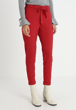 Pantalon rouge avec une ceinture à nouer à la taille, coupe slim, ourlets retournés et tissu lisse. Associé à des bottines de couleur claire.