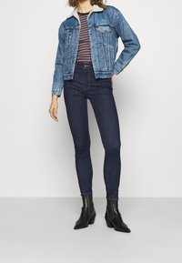 Chaqueta de mezclilla con collar de borreguito, color azul claro, bolsillos frontales; camiseta a rayas; jeans oscuros ajustados; botas negras hasta el tobillo con tacón cuadrado.