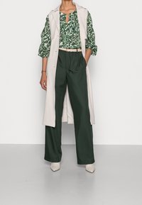 Une femme porte un chemisier floral vert, un manteau sans manches beige, un pantalon large vert foncé, une ceinture beige et des bottes à bout pointu beiges, se tenant avec une main dans la poche.