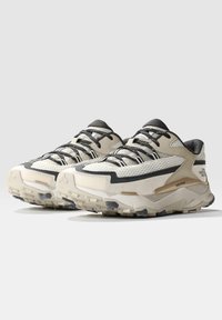 Zapatos deportivos en beige claro y gris, con materiales de malla y gamuza, suelas texturizadas y un diseño con cordones que presenta un branding mínimo.