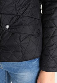 Casaco preto acolchoado com fecho de pressão, logótipo bordado e padrão de costura diagonal. Modelo a usar jeans azuis claros.