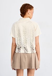 Kortärmad krämfärgad polo med ett leopardmönster i gradient vid ryggen, tillverkad av ett strukturerat tyg, kombinerad med en beige veckad kjol.
