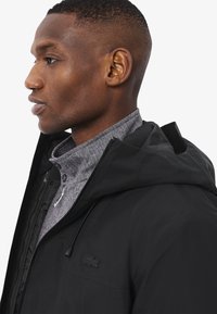 Profilo di un uomo che indossa una giacca nera con cappuccio sopra un maglione grigio con zip e motivo a spina di pesce, rivolto verso sinistra.