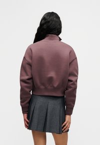 Σκούρο μοβ κοντό φούτερ με όρθιο γιακά και χαλαρά ραμμένα ώμους, σε συνδυασμό με γκρι πλισέ φούστα. Απαλή υφή υφάσματος.