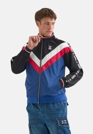 Blue track jacket with black and red chevron accents, white stripes, zip front, ribbed collar, and logo on the chest and sleeves.