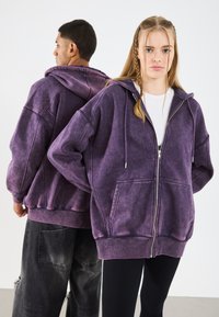 Felpa con zip color viola in pile, dalla vestibilità ampia, con tasche frontali e un grande cappuccio, caratterizzata da una texture spazzolata e leggere variazioni di colore.