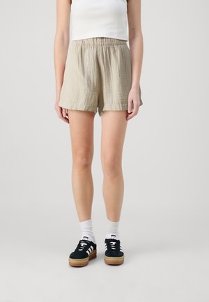 Person trägt beige, strukturierte Shorts, weißes ärmelloses Oberteil, weiße Socken und schwarze Sneakers mit weißen Streifen und steht vor einem schlichten Hintergrund.