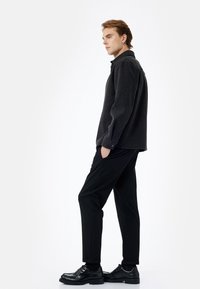 Mørk grå sweatshirt med krave og lange ærmer, parret med sorte tapered bukser. Modellen har sorte snørede sko på. Enkle, rene linjer gennem hele designet.