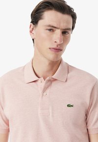 Ljusrosa polo-shirt med krage, tre knappar och en liten broderad grön krokodil-logotyp på vänster bröst. Material i bomull med textur.