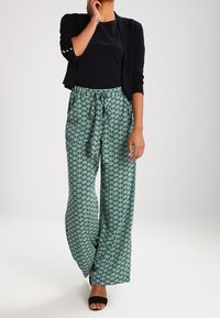 Pantalon à jambes larges en sarcelle avec un motif géométrique, associé à un haut noir et un blazer. Il présente une taille nouée et un tissu lisse.