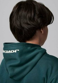 Felpa verde scuro con una texture morbida, dotata di un ampio cappuccio e logo bianco sul retro. Focalizzata sui dettagli del tessuto e del design.