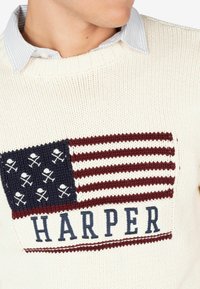 Suéter de punto color crema con un diseño de bandera americana tejido en azul marino y rojo, adornado con calaveras. "HARPER" impreso debajo de la bandera.
