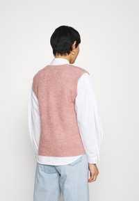 Gilet rosa in maglia con texture a coste, design senza maniche, scollatura rotonda, indossato sopra una camicia bianca a maniche lunghe, vista posteriore.