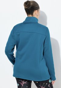 Tyrkysová fleecová bunda s vysokým límcem, s hladkou texturou a přiléhavým střihem, v kombinaci s legínami s nebeskými motivy.