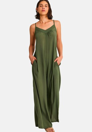 Femme portant une robe longue olive sans manches avec un col en V et des poches, debout les mains dans les poches devant un fond uni.