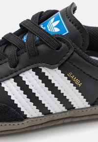 Svarta lädersportskor med vita zigzag-ränder, texturerat svart mocka-akcent, blå logotyp-patch och guld "SAMBA"-detalj på sidan.