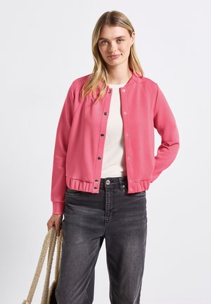 Veste bombers rose avec des boutons, des poignets et un ourlet côtelés. Portée par-dessus une chemise crème, associée à un jean gris. Modèle tenant un sac en osier.