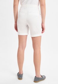 Weiße Baumwollshorts mit umgeschlagenen Säumen, zwei Gesäßtaschen und hoher Taille. Getragen mit grauen Slippern. Glatte Textur, lässiges Design.