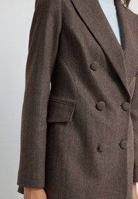 Cappotto doppiopetto marrone a motivo spigato con rever ad angolo, tasche frontali e tessuto testurizzato. Presenta bottoni tondi e vestibilità sartoriale.