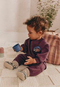 Lilla bomuldsoutfit med en rund applikation, der viser en bjørn. Strikkede sokker med geometrisk mønster. Barnet holder en blå kop. Tekstureret gulvmåtte.