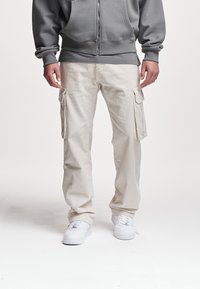 Beige kordfløyels cargo-bukser med store sidelommer, avslappet passform og teksturert overflate. Kombinert med hvite joggesko og en grå hettegenser.