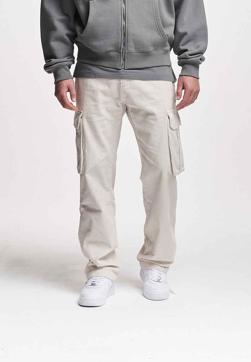 Beige kordfløyels cargo-bukser med store sidelommer, avslappet passform og teksturert overflate. Kombinert med hvite joggesko og en grå hettegenser.