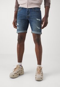 Denimshorts in mittlerem Waschton mit umgeschlagenen Säumen und distressed Details. Getragen mit beigen, atmungsaktiven Schuhen und gestreiften Knöchelsocken.