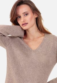 Pull en tricot beige avec un col en V, des manches longues et des accents texturés à l'épaule. Matière douce et confortable avec une coupe décontractée.