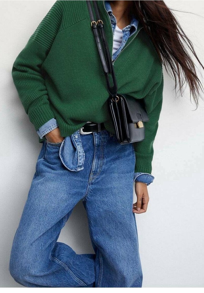 Femme portant un pull en laine vert par-dessus une chemise en jean rentrée dans un jean bleu taille haute avec une ceinture noire et un sac noir porté en bandoulière.