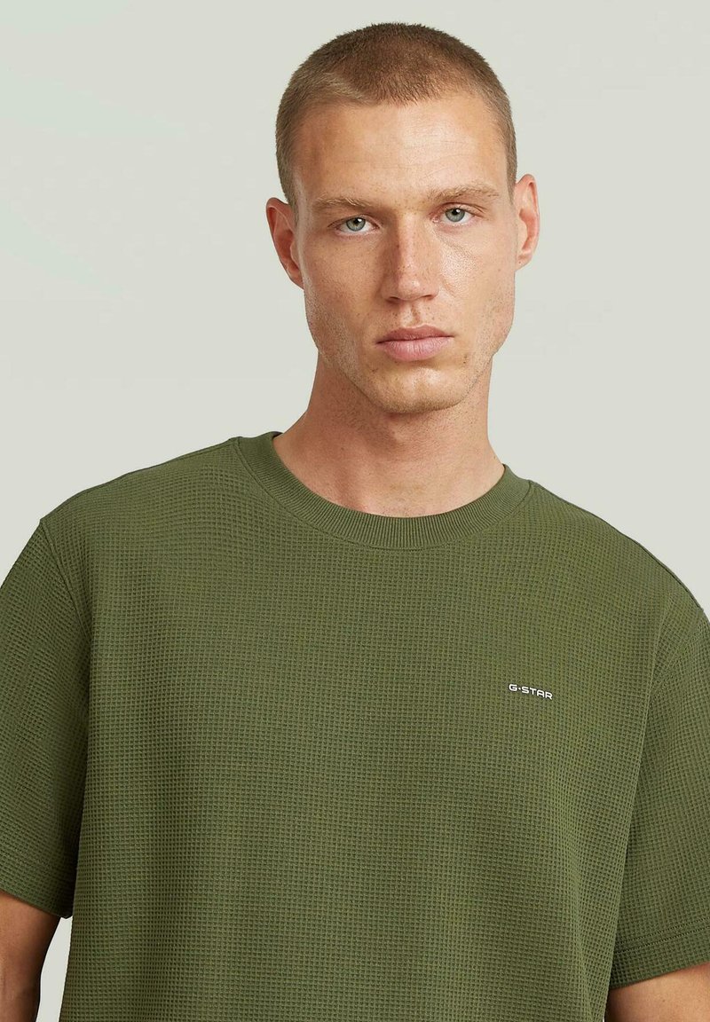Hombre joven con cabello corto, vistiendo una camiseta texturizada de G-Star color verde oliva, mirando hacia adelante con una expresión neutral.