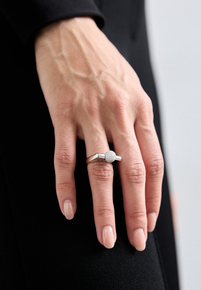 Sudraba gredzens ar teksturētu apaļu centri un gludu joslu, kas uzlikts uz rokas ar gaišām, pulētām nagu nagiem uz tumša fona.