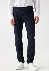 Jeans de mezclilla azul oscuro con un corte slim, que cuentan con cinco bolsillos, costuras naranjas y un cierre de botón en la cintura. Combinados con zapatillas blancas.