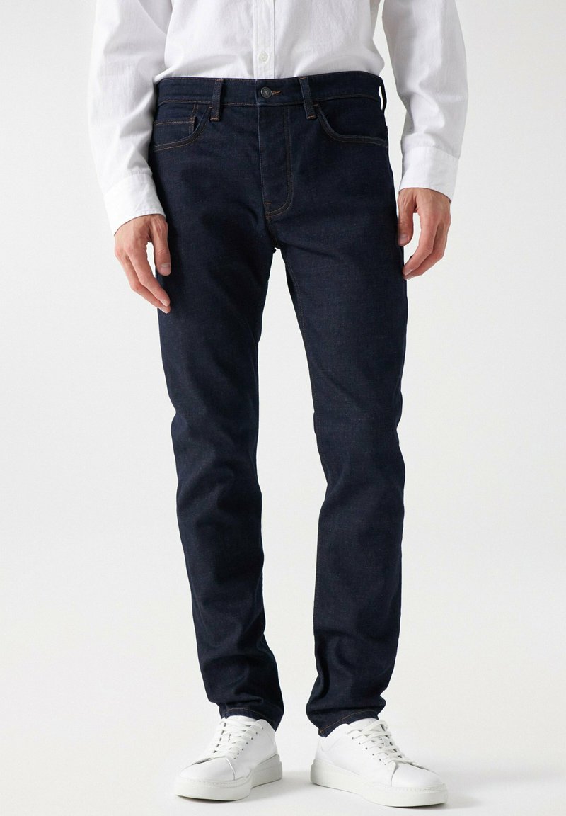 Jeans de mezclilla azul oscuro con un corte slim, que cuentan con cinco bolsillos, costuras naranjas y un cierre de botón en la cintura. Combinados con zapatillas blancas.