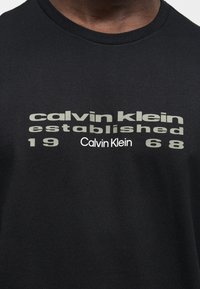 Gros plan d'un T-shirt noir avec un texte gris fané "calvin klein established 1968" et un plus petit logo blanc "Calvin Klein" en dessous.