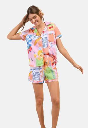 Young woman smiling, wearing colorful short-sleeve button-up shirt and shorts with fruit and food illustrations, standing with one arm raised.