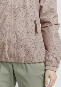 Giacca rosa chiaro a fantasia con pois blu navy, chiusura con zip, polsini elasticizzati e dettagli con tasche laterali. Abbinata a pantaloni verdi.