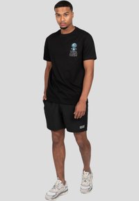 Camiseta de algodón negra con gráfica y texto azules en el pecho, combinada con pantalones cortos negros. El modelo lleva zapatillas deportivas grises con acentos blancos.