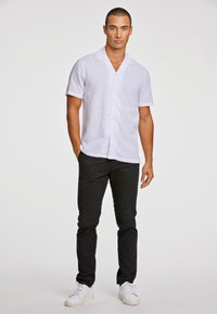 Camisa de lino blanca de manga corta con cuello con muesca, combinada con pantalones negros y zapatillas blancas. La camisa tiene un cierre con botones en la parte delantera.