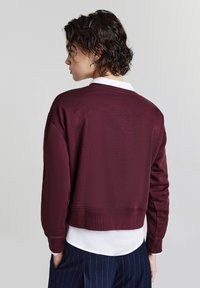 Persona con capelli corti e ricci che indossa un maglione bordeaux sopra una camicia bianca e pantaloni navy a righe, in posizione laterale con le mani nelle tasche.