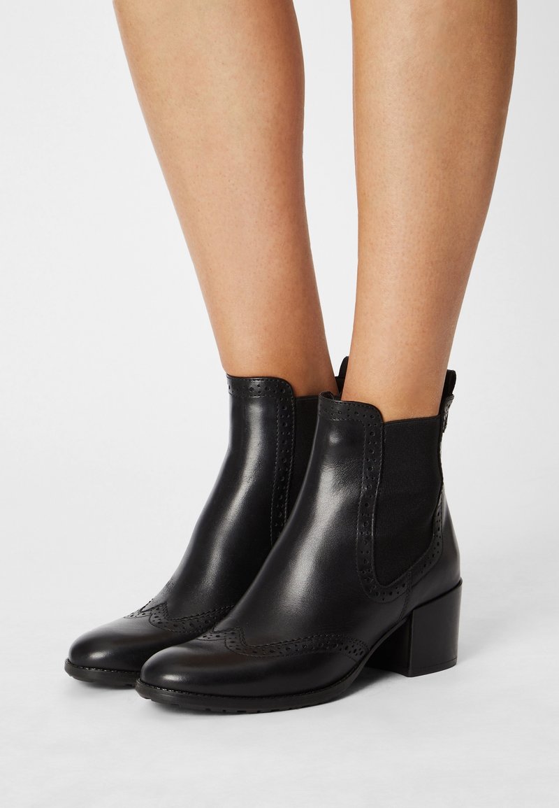 Bottines en cuir noir avec des détails en brogue et des talons blocs, portées sur des jambes nues contre un fond clair.