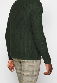 Homme portant un pull en tricot texturé vert foncé et un pantalon à carreaux gris, vu de dos sur un fond uni.